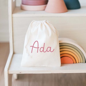 May include: A white wooden step stool with two steps. On the top step is a white drawstring bag with the name 'Ada' embroidered on it in pink. There is a rainbow wooden toy and a stack of colorful wooden rings on the lower step.