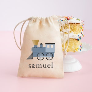 Könnte beinhalten: Eine kleine, beige Kordeltasche mit einem blauen Zugmotiv und dem Namen "samuel" auf der Vorderseite. Die Tasche liegt auf einer rosa Oberfläche neben einem weißen Kuchen mit bunten Streuseln.