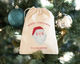 Puede incluir: Una bolsa de regalo de tela blanca con una ilustración de Papá Noel roja y blanca y el texto "Feliz Navidad para: Harper". La bolsa está colgada de un árbol de Navidad verde con adornos.