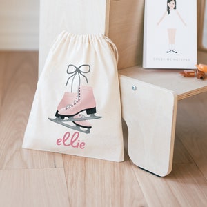 Puede incluir: Una bolsa de tela blanca con cordón de ajuste con un diseño de patines de hielo rosas y el nombre "ellie" impreso en la parte delantera. La bolsa está sentada en un taburete de madera con una cámara azul y un bloc de notas para vestir en el escalón superior.