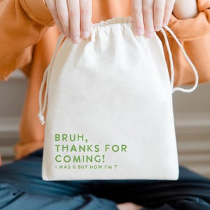 May include: A cream-coloured drawstring bag with green text saying "BRUH, THANKS FOR COMING! I WAS 6 BUT NOW I'M 7." The bag is held by someone wearing an orange long-sleeved top and dark blue trousers. The bag is likely a party favour.
