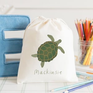 May include: A white drawstring bag with a green sea turtle illustration and the name "Mackinsie" printed on the front. The bag is sitting on a table with a blue corduroy letter E and a jar of colored pencils.