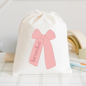 Puede incluir: Una bolsa de tela blanca con un lazo rosa y el nombre "Brooke" impreso en ella.