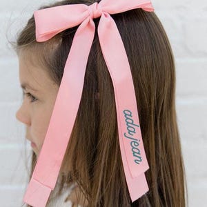 May include: A pale pink bow hair accessory with long ribbons. The bow is tied at the top of the head. The text "ada jean" is printed on one of the ribbons. The girl is wearing a white dress with pink flower embroidery.