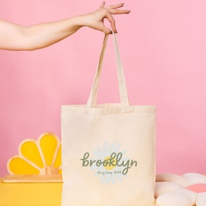 Pode incluir: Uma bolsa de lona branca com uma margarida e o texto "brooklyn daisy troop #476" impresso nela.