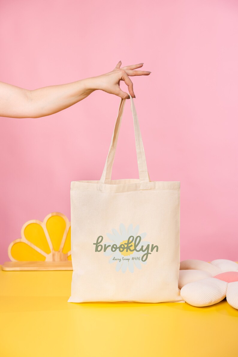 Pode incluir: Uma bolsa de lona branca com uma margarida e o texto "brooklyn daisy troop #476" impresso nela.