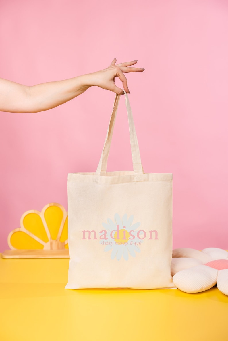 Pode incluir: Uma bolsa tote branca com um desenho de margarida e o texto "madison daisy troop #476".