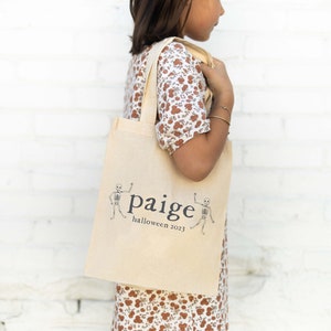 Puede incluir: Un bolso tote de lona beige con el nombre "Paige", el texto "halloween 2023" e ilustraciones de esqueletos. El bolso tiene dos asas y lo sostiene una persona. El diseño es sencillo y adecuado para Halloween.