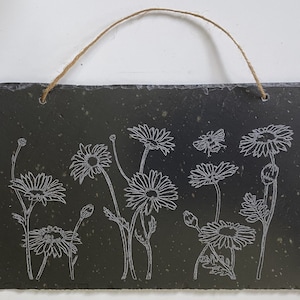 May include: Black slate hanging sign with a white design of daisies and a bee. The sign has a rope hanger.