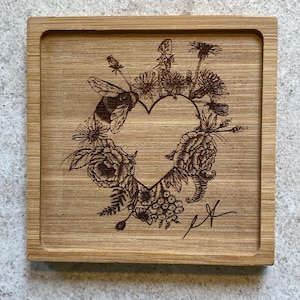 May include: A wooden coaster with a laser-engraved design of a heart surrounded by flowers and a bee. The heart is formed by the flowers and leaves. The coaster has a natural wood grain finish.
