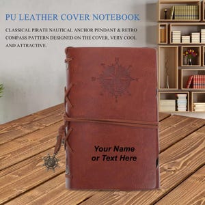 Puede incluir: Cuaderno de cuero marrón con un diseño de brújula y ancla. El cuaderno tiene un cierre de correa de cuero y un colgante de ancla de metal. El texto "Your Name or Text Here" está grabado en la portada.