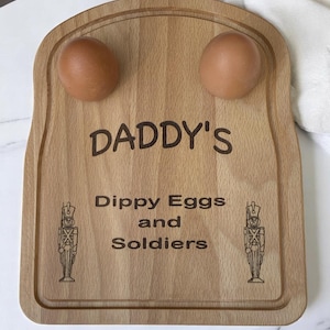 Puede incluir: Tabla de desayuno de madera con dos huevos marrones. La tabla está grabada con las palabras "DADDY'S Dippy Eggs and Soldiers" y presenta ilustraciones de soldados. La tabla es de color marrón claro.