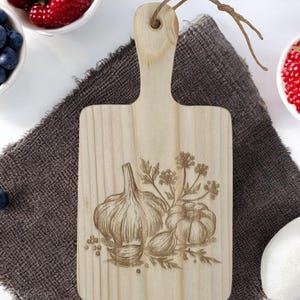 Può includere: Un piccolo tagliere di legno con un manico. Il tagliere ha un disegno di aglio ed erbe intagliato sulla superficie.