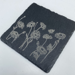 May include: Square, dark gray slate coaster with a floral design. The design features etched outlines of daisies and a bee. The coaster has a rough, natural edge and is suitable for use with beverages.