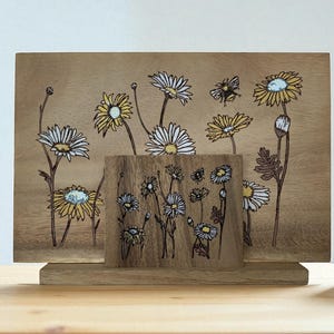 Op de afbeelding: Houten kunstwerk met een madeliefjes bloemenontwerp. Het kunstwerk bestaat uit een rechthoekig houten paneel en een kleiner, vierkant houten stuk ervoor. De madeliefjes hebben witte bloemblaadjes en gele centra.