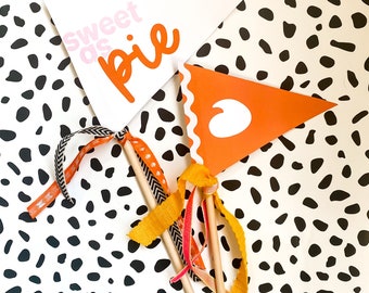 May include: Two triangular pennants on wooden sticks with colorful ribbons. One white pennant has the text "sweet as pie" in pink and orange. The other is orange with a white heart shape and scalloped edge. The background is white with black spots.