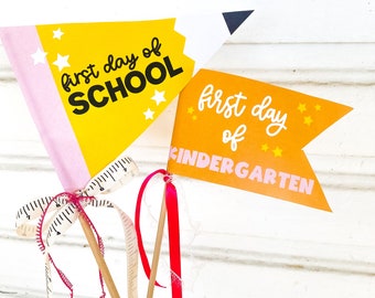 Op de afbeelding: Schoolvlaggen met de zinnen "first day of SCHOOL" en "first day of KINDERGARTEN" in witte letters. De vlaggen zijn geel en oranje met steraccenten. De vlaggen zijn bevestigd aan houten deuvels met roze en rode linten.