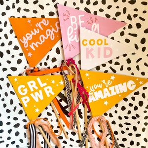 May include: A set of ten colourful pennant flags with positive messages, including "You're Magic", "Be Kind", "Cool Kid", "Grl Pwr", and "You're Amazing". The flags are on wooden sticks and have a white polka dot background.