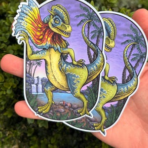 Può includere: Due adesivi fustellati raffiguranti un dinosauro Dilophosaurus con un corpo giallo e blu vivace e un grande collare colorato rosso, arancione e blu. Lo sfondo mostra un paesaggio tropicale con palme, una cascata e un lago.
