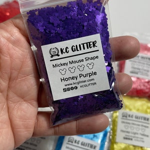 May include: A small clear plastic bag filled with purple glitter shaped like Mickey Mouse heads. The bag has a white label with the text "KC GLITTER", "Mickey Mouse Shape", "Honey Purple", "www.kcglitter.com", and "KCGLITTER".