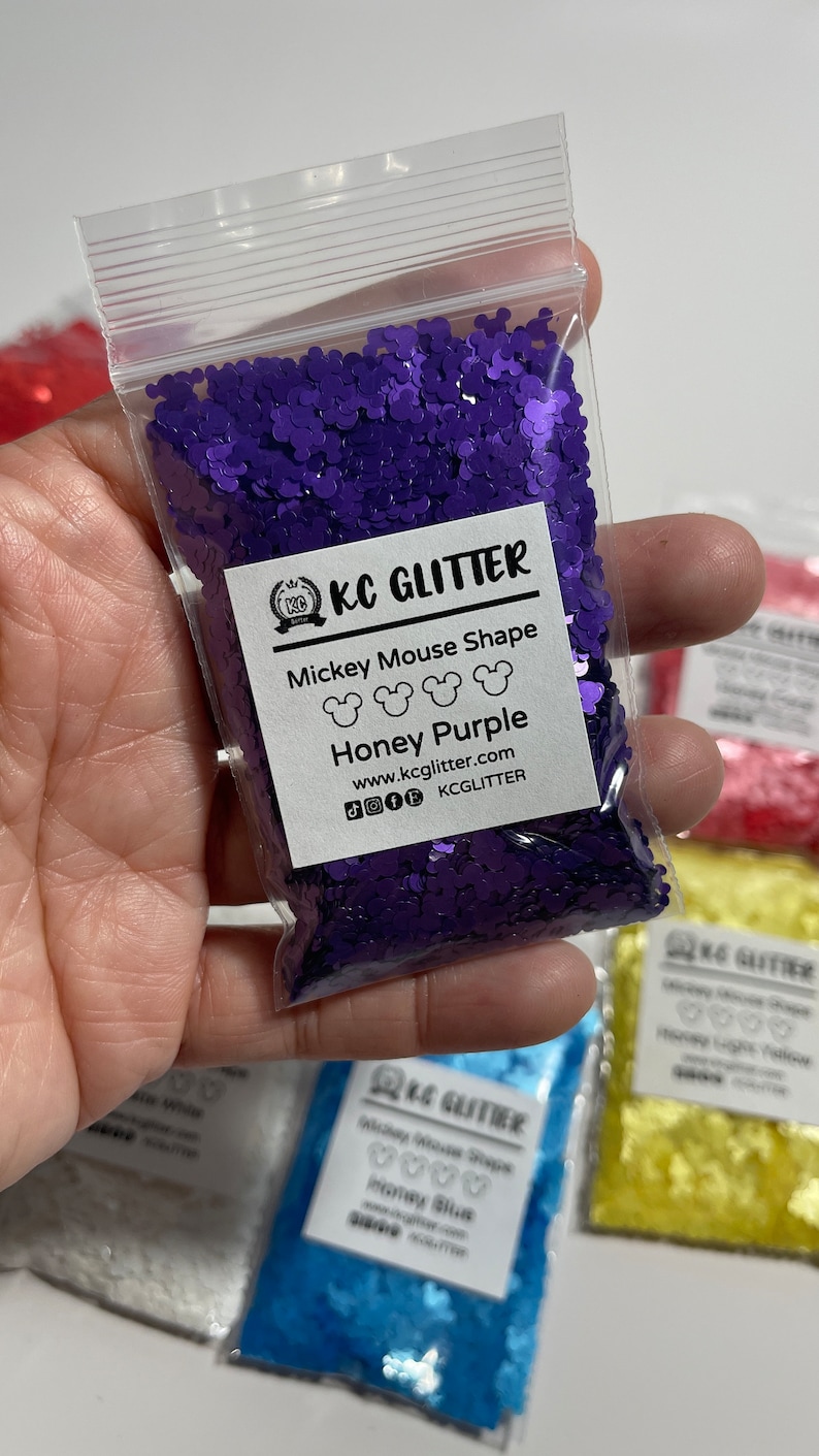 May include: A small clear plastic bag filled with purple glitter shaped like Mickey Mouse heads. The bag has a white label with the text "KC GLITTER", "Mickey Mouse Shape", "Honey Purple", "www.kcglitter.com", and "KCGLITTER".