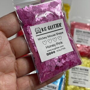 May include: A small plastic bag filled with pink glitter shaped like Mickey Mouse heads. The bag has a white label with the text "KC GLITTER", "Mickey Mouse Shape", "Honey Pink", "www.kcglitter.com", and "KCGLITTER".