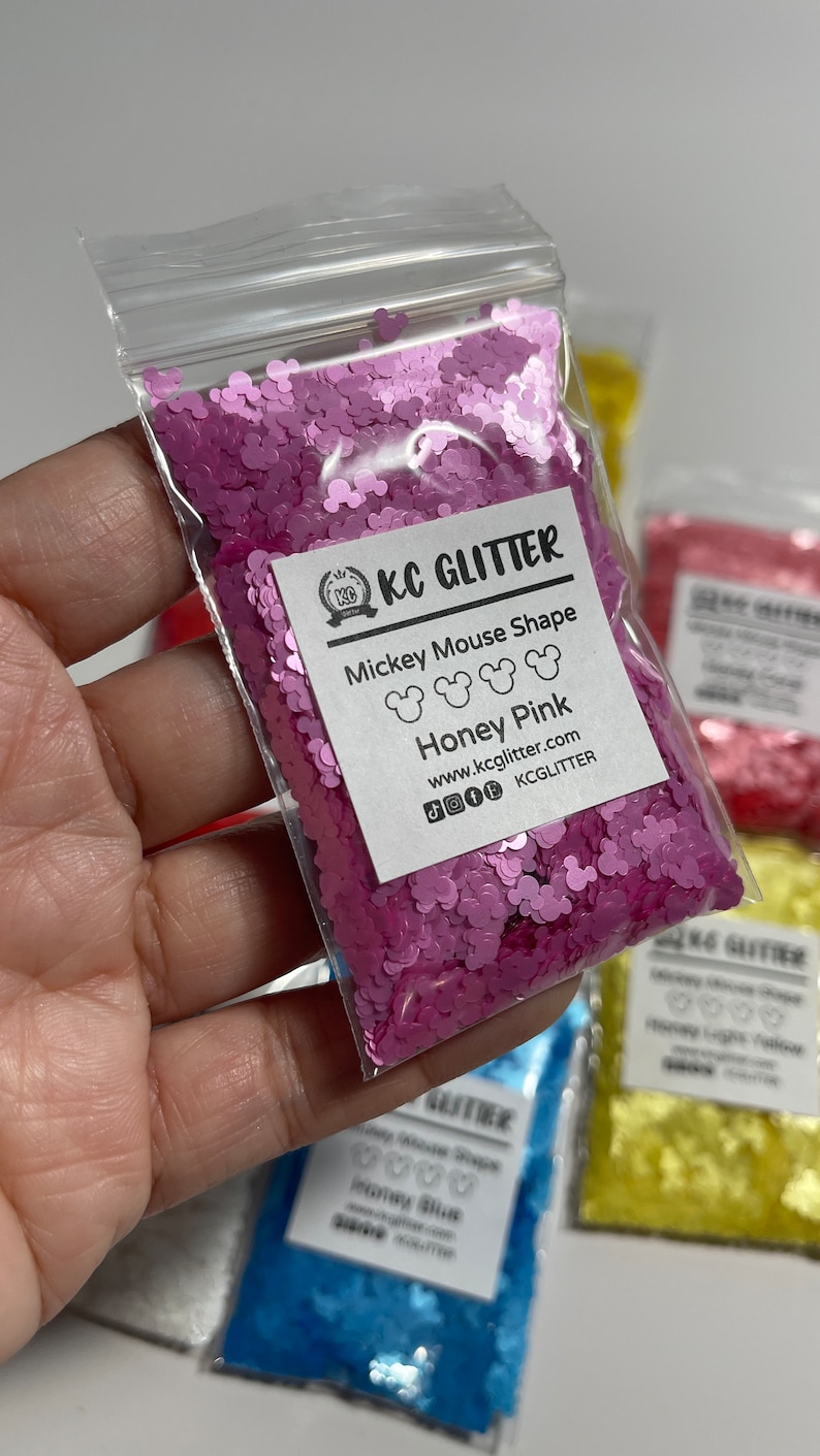 May include: A small plastic bag filled with pink glitter shaped like Mickey Mouse heads. The bag has a white label with the text "KC GLITTER", "Mickey Mouse Shape", "Honey Pink", "www.kcglitter.com", and "KCGLITTER".