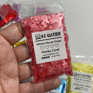 May include: A small plastic bag filled with pink glitter shaped like Mickey Mouse heads. The bag has a label that reads "KC GLITTER" and "Mickey Mouse Shape Honey Coral".