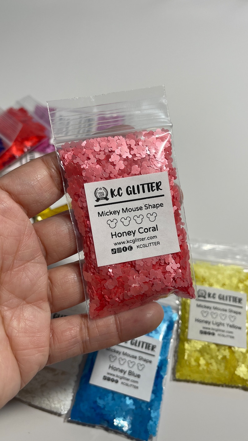 May include: A small plastic bag filled with pink glitter shaped like Mickey Mouse heads. The bag has a label that reads "KC GLITTER" and "Mickey Mouse Shape Honey Coral".