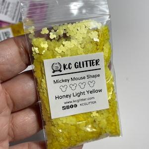 May include: A small clear plastic bag filled with yellow glitter shaped like Mickey Mouse heads. The bag has a white label that reads "KC GLITTER", "Mickey Mouse Shape", "Honey Light Yellow", "www.kcglitter.com", and "KCGlitter".