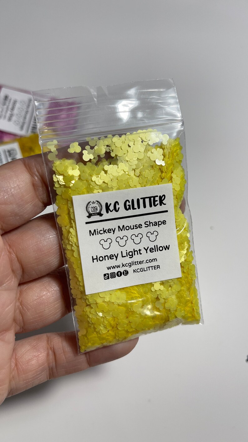 May include: A small clear plastic bag filled with yellow glitter shaped like Mickey Mouse heads. The bag has a white label that reads "KC GLITTER", "Mickey Mouse Shape", "Honey Light Yellow", "www.kcglitter.com", and "KCGlitter".