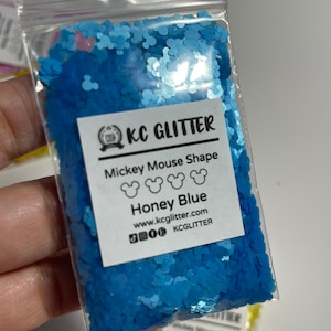May include: A small plastic bag filled with blue glitter shaped like Mickey Mouse heads. The bag has a label that says "KC Glitter" and "Mickey Mouse Shape Honey Blue". The label also includes the website address "www.kcglitter.com" and the social media handle "KCGlitter".