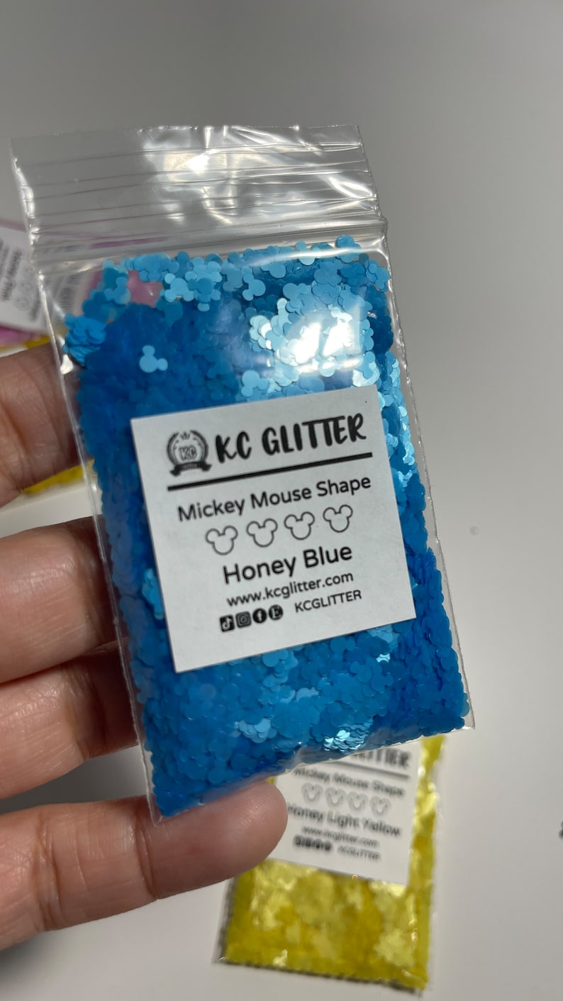 May include: A small plastic bag filled with blue glitter shaped like Mickey Mouse heads. The bag has a label that says "KC Glitter" and "Mickey Mouse Shape Honey Blue". The label also includes the website address "www.kcglitter.com" and the social media handle "KCGlitter".