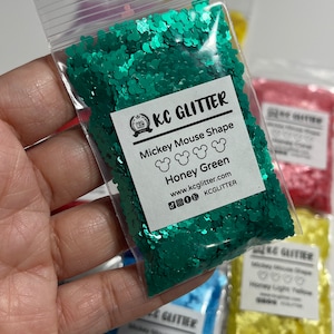 May include: A close-up of a small plastic bag filled with green glitter shaped like Mickey Mouse heads. The bag has a label that reads "KC Glitter Mickey Mouse Shape Honey Green www.kcglitter.com KCGLITTER".