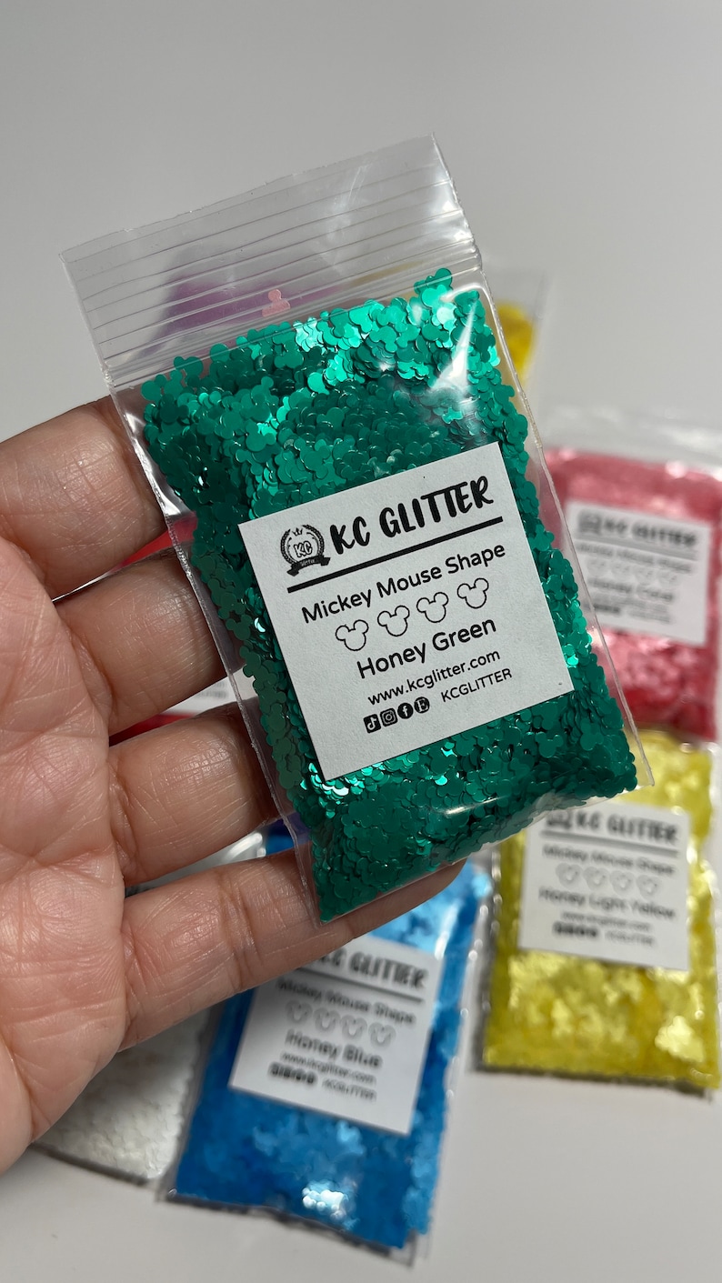 May include: A close-up of a small plastic bag filled with green glitter shaped like Mickey Mouse heads. The bag has a label that reads "KC Glitter Mickey Mouse Shape Honey Green www.kcglitter.com KCGLITTER".