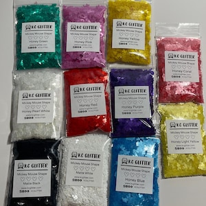 May include: Ten small bags of glitter shaped like Mickey Mouse heads. Each bag has a different color glitter and a label with the color name and the company name "KC Glitter".