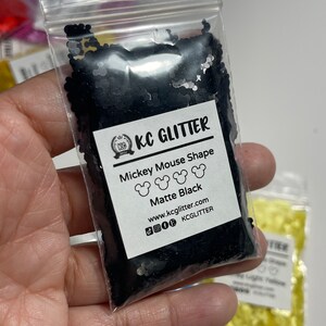 May include: A small clear plastic bag filled with black glitter shaped like Mickey Mouse heads. The bag has a white label that says "KC GLITTER", "Mickey Mouse Shape", "Matte Black", "www.kcglitter.com", and "KCGLITTER".