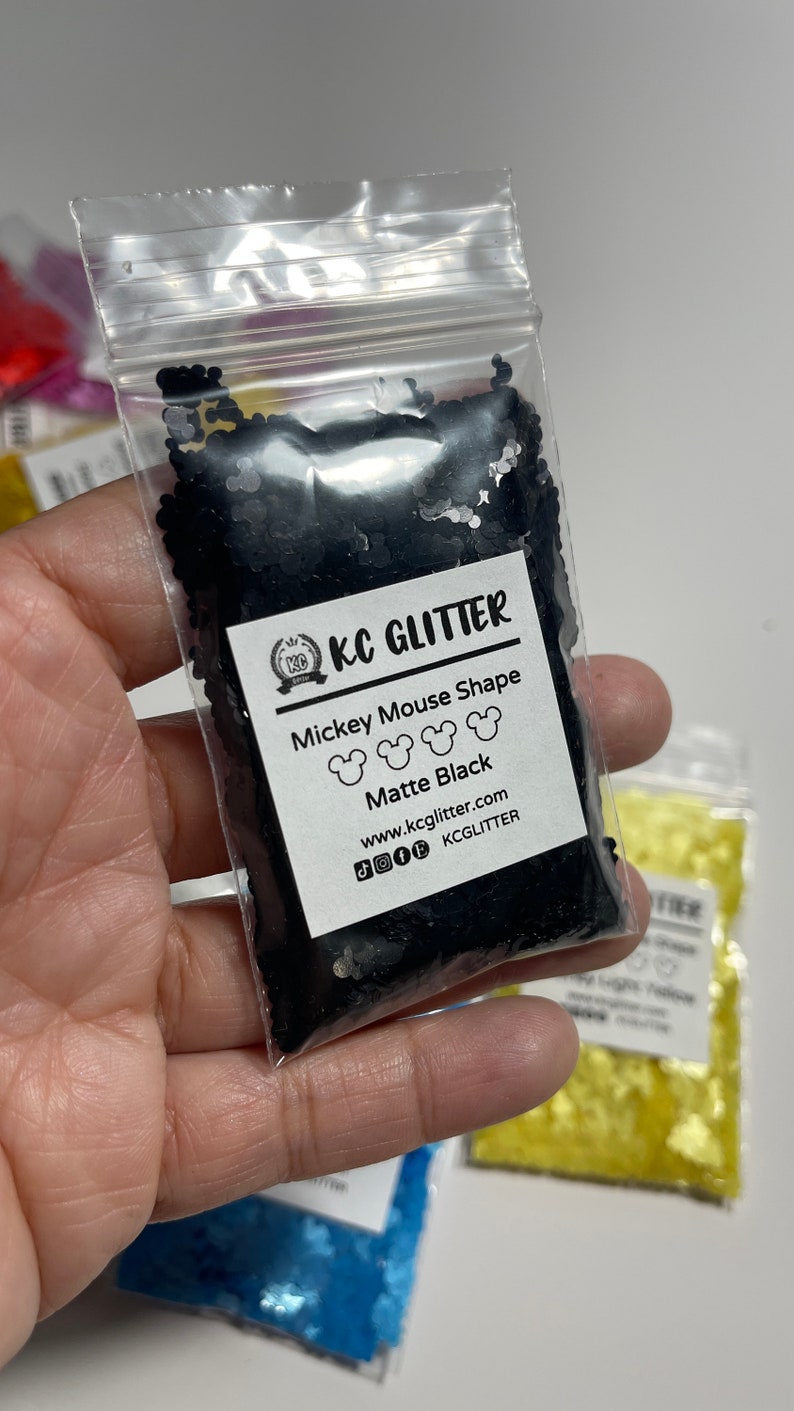 May include: A small clear plastic bag filled with black glitter shaped like Mickey Mouse heads. The bag has a white label that says "KC GLITTER", "Mickey Mouse Shape", "Matte Black", "www.kcglitter.com", and "KCGLITTER".