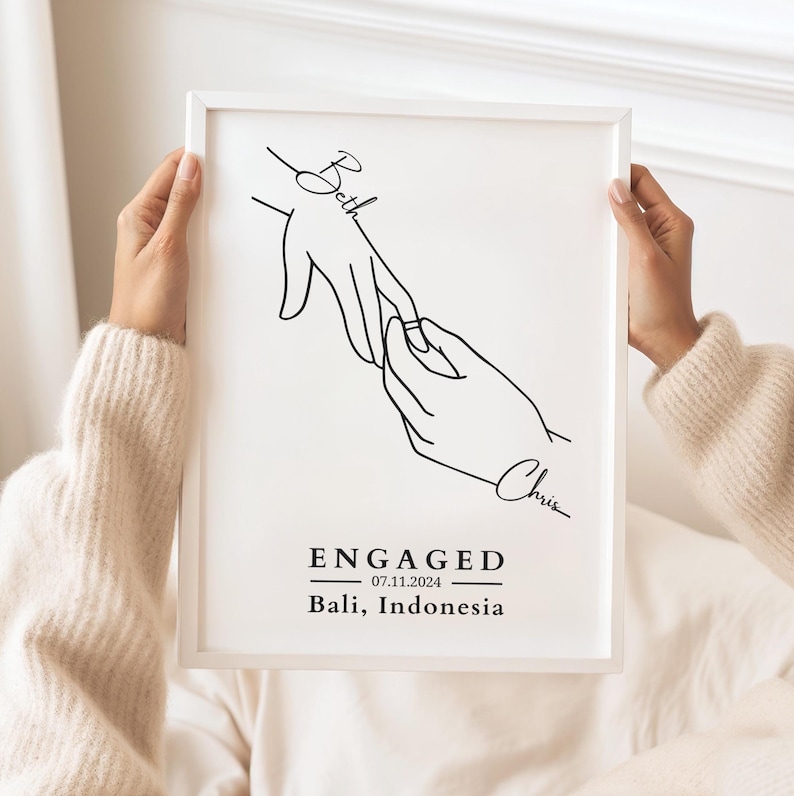 May include: A framed black and white line art print featuring two hands, one placing a ring on the other's finger. The word "Beth" is written above one hand, and "Chris" below the other. The word "ENGAGED" is printed below, along with the date and location.