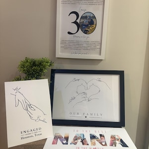 a display of various framed artworks and a sign, including a framed quote, a framed drawing of hands, and a sign that reads "To the best NANA in the world".