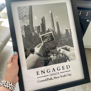 May include: Black and white framed print with the word "ENGAGED" and the date 29/03/24, along with "Central Park, New York City". The image shows a cityscape and a sonogram. A hand holds the frame.