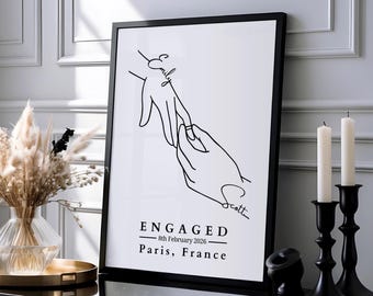 May include: A black and white framed print featuring a line art illustration of two hands, one placing a ring on the other. The names Emily and Scott are written in script. Text reads: ENGAGED, 8th February 2026, Paris, France.
