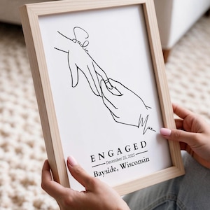 May include: A framed print with a minimalist line drawing of two hands, one with a ring being placed on a finger. The print includes the word "ENGAGED", the date December 25, 2025, and the location Bayside, Wisconsin.