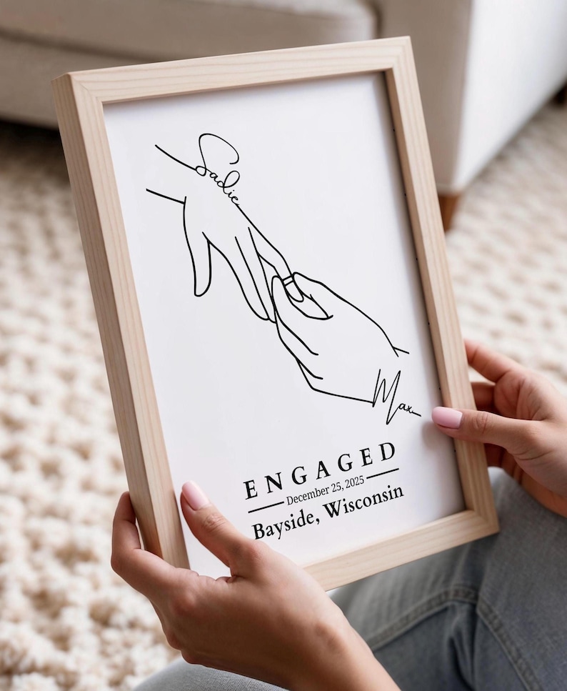 May include: A framed print with a minimalist line drawing of two hands, one with a ring being placed on a finger. The print includes the word "ENGAGED", the date December 25, 2025, and the location Bayside, Wisconsin.