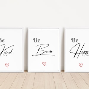 Op de afbeelding: Drie ingelijste kunstdrukken met witte achtergronden. Elke print bevat het woord "Be" in grijs, gevolgd door een cursief woord in zwart: "Kind", "Brave" en "Happy". Onder elk woord staat een klein rood hart.
