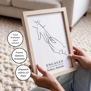 May include: A framed print with a minimalist line drawing of two hands, one placing a ring on the other. The print includes the names "Hannah" and "Charlie", and the word "ENGAGED" with the date and location.