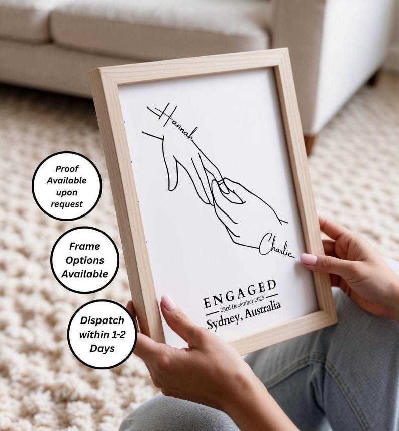 May include: A framed print with a minimalist line drawing of two hands, one placing a ring on the other. The print includes the names "Hannah" and "Charlie", and the word "ENGAGED" with the date and location.