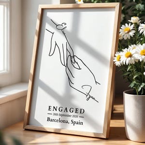 Framed engagement print on a sunlit wooden table beside a window and white daisies. The minimalist black line drawing shows one hand placing a ring on another, with the names “Elisa” and “Flynn.” Below, the text reads: “ENGAGED — 26th September 2026 — Barcelona, Spain.”