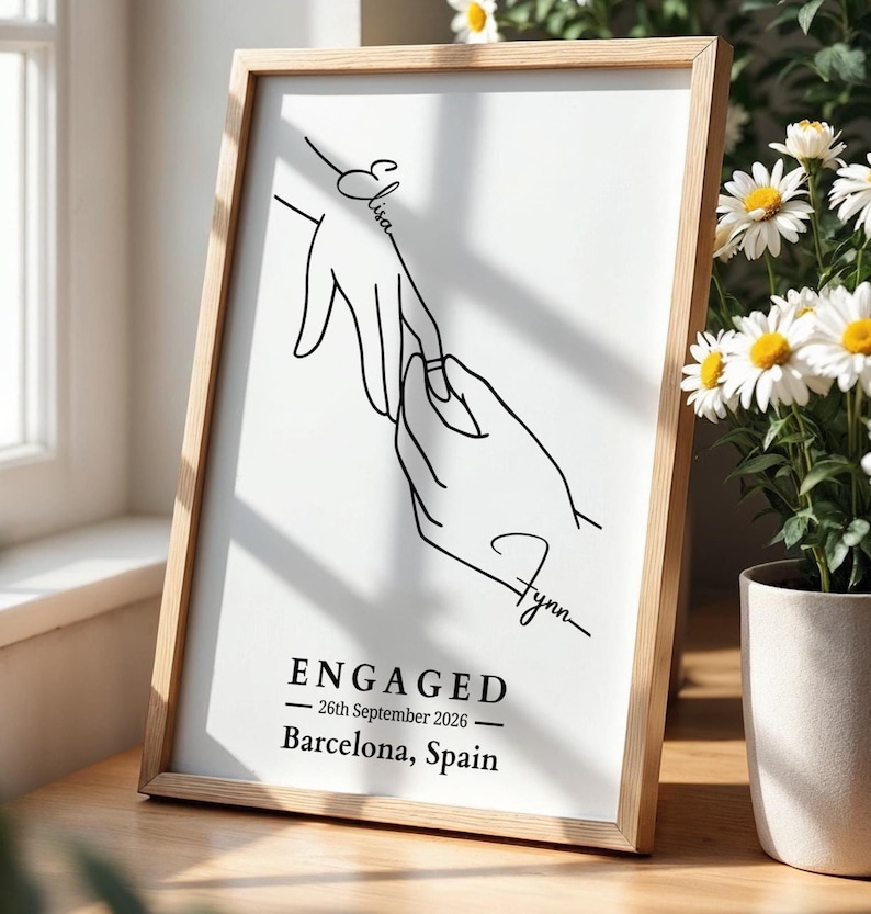 Framed engagement print on a sunlit wooden table beside a window and white daisies. The minimalist black line drawing shows one hand placing a ring on another, with the names “Elisa” and “Flynn.” Below, the text reads: “ENGAGED — 26th September 2026 — Barcelona, Spain.”