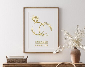 May include: A framed wall print featuring a gold-colored graphic of two interlocking rings, one topped with a diamond. The text reads Emily, Christopher, ENGAGED, 09/05/2024, and London, UK, all displayed in a gold font on a white background.